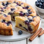 Slice of blueberry breakfast cake on a white plate with fresh blueberries and cinnamon sticks