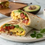 andscape view of a breakfast burrito cut in half and stacked, revealing eggs, bacon, melted cheddar, avocado, tomato, and salsa on a white plate.
